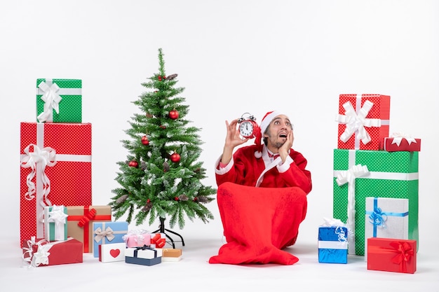 Emotionaler glücklicher überraschter Weihnachtsmann, der oben schaut auf dem Boden sitzt und Uhr nahe Geschenken und geschmücktem Weihnachtsbaum auf weißem Hintergrund zeigt