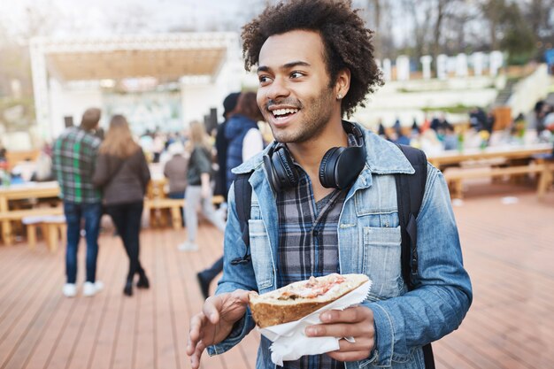 Emotionaler gutaussehender dunkelhäutiger Mann mit Borste, der Sandwich hält, während er gestikuliert und Gedanken oder Eindrücke mit Freunden teilt, auf Food Festival oder Konzert im Park ist