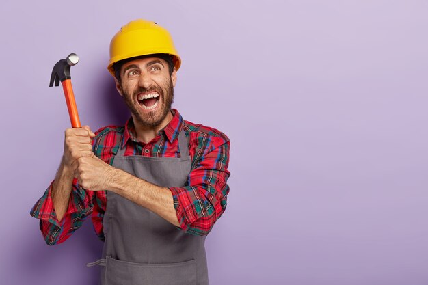 Emotionaler Handwerker hält Hammer mit beiden Händen, bereit für die Arbeit, trägt gelben Schutzhelm, Freizeitarbeitskleidung, schreit emotional