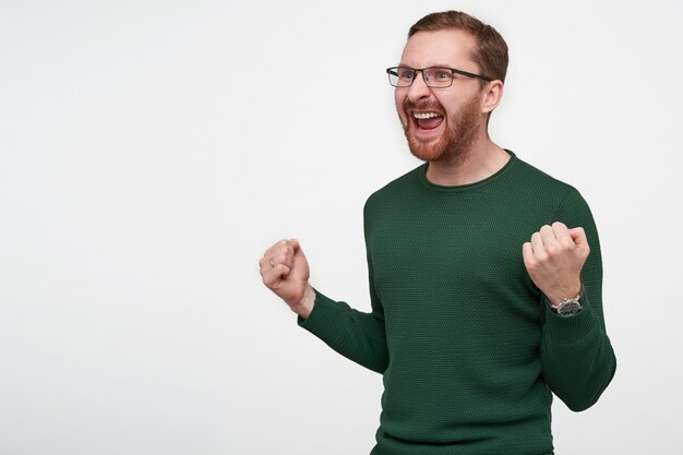Emotionaler junger bärtiger mann in brille mit braunen kurzen haaren, die aufgeregt zur seite schauen, mit weit geöffnetem mund und erhobenen händen, posierend