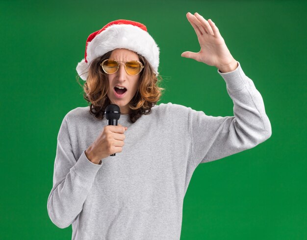 Emotionaler junger Mann, der Weihnachtsweihnachtsmütze und gelbe Brille trägt, die zum Mikrofon mit erhöhtem Arm schreien, der über grünem Hintergrund steht