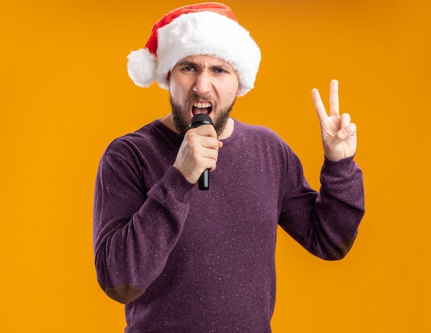 Emotionaler junger Mann im lila Pullover und in der Weihnachtsmannmütze, die Mikrofon hält, das v-Zeichen singend steht über orange Hintergrund
