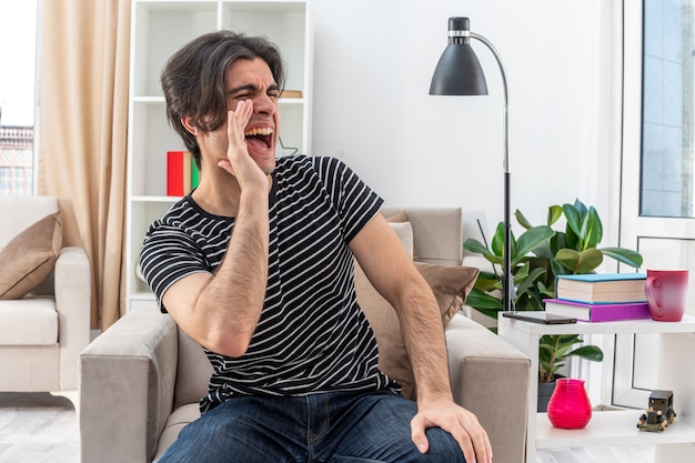 Emotionaler junger Mann in Freizeitkleidung, der laut schreit oder jemanden mit der Hand in der Nähe des Mundes anruft, der auf dem Stuhl im hellen Wohnzimmer sitzt