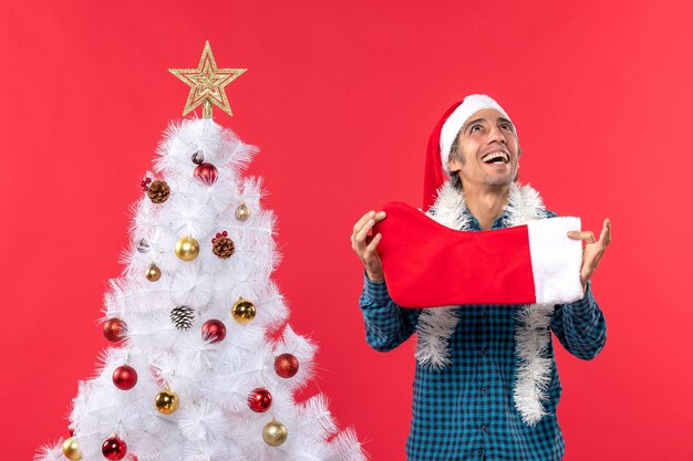 Emotionaler junger Mann mit Weihnachtsmannhut in einem blauen gestreiften Hemd und Halten der Weihnachtssocke, die oben schaut