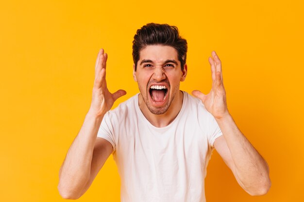 Emotionaler Mann im weißen T-Shirt schreit wütend und schaut in die Kamera auf orange Raum.