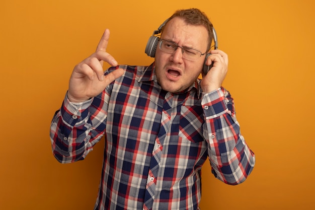 Emotionaler Mann in der Brille und im karierten Hemd mit den Kopfhörern, die zur Seite schauen und Zeigefinger stehen, der über orange Wand steht