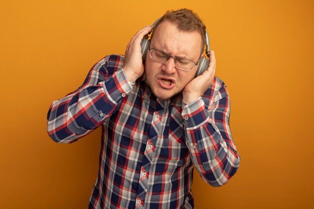 Emotionaler Mann in der Brille und im karierten Hemd mit Kopfhörern, die Musik genießen, die über orange Wand steht