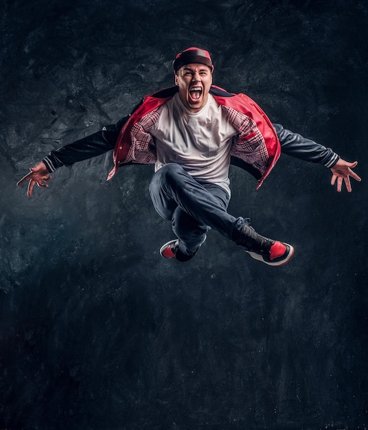 Emotionaler, stilvoll gekleideter Typ, der Breakdance-Springen durchführt. Studiofoto gegen eine dunkle strukturierte Wand