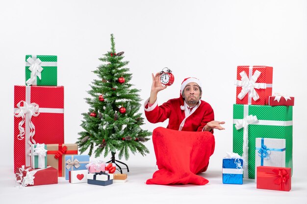 Emotionaler verrückter überraschter verwirrter Weihnachtsmann, der auf dem Boden sitzt und Uhr nahe Geschenken und geschmücktem Weihnachtsbaum auf weißem Hintergrund zeigt