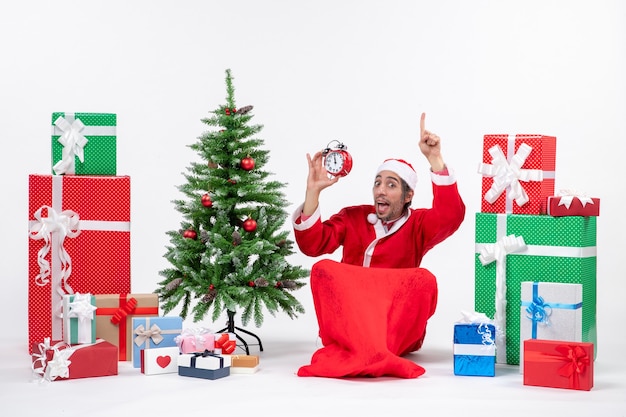 Emotionaler verrückter überraschter weihnachtsmann, der auf dem boden sitzt und uhr zeigt, die oben nahe geschenken und geschmücktem weihnachtsbaum auf weißem hintergrund zeigt