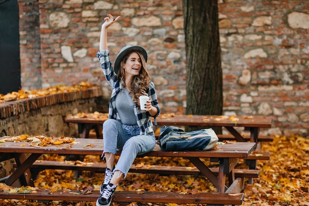 Emotionales weibliches Modell in kurzen Jeans und in schwarzen Schuhen trinkt Kaffee im Park im warmen Septembermorgen