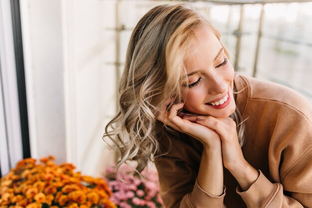 Emotionales weißes Mädchen, das mit geschlossenen Augen lächelt. Porträt der begeisterten lockigen Frau, die nahe orange Blumen sitzt.