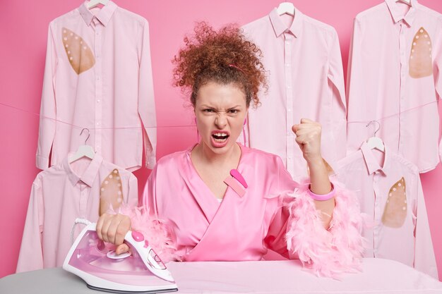 Empört verärgert Hausfrau hassen Bügeln ballt Fäuste schreit wütend hat lockiges Haar in häuslicher Kleidung gekleidet. Gereizter Wäschereiarbeiter bügelt Hemden im Reinigungssalon. Housekeeping-Konzept