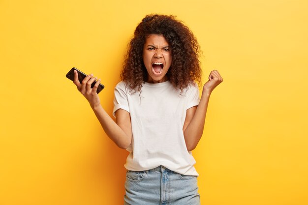 Empörte junge Frau mit lockigem Haar posiert mit ihrem Telefon