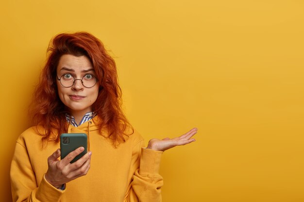 Empörte verwirrte rothaarige Frau hebt die Handfläche, überlegt, was sie auf die empfangene Nachricht antworten soll, hält das Handy in der Hand, trägt eine runde Brille und einen Kapuzenpulli, Modelle über der gelben Wand mit Leerzeichen rechts.