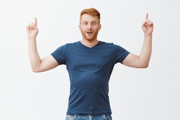 Endlich die Spitze des Erfolgs erreichen, kann es nicht glauben. Porträt einer beeindruckten aufgeregten und verblüfften hübschen erwachsenen Rothaarigen mit Borsten im blauen T-Shirt, die mit erhobenen Händen nach oben zeigt, entzückt und erstaunt