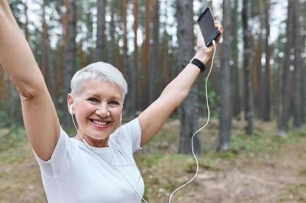 Energetische fröhliche pensionierte Frau mit fit schlankem Körper, der draußen in Kopfhörern aufwirft, Hände hebt, Handy hält