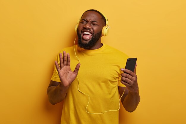 Energetischer afroamerikanischer Mann zieht Handfläche in Richtung Kamera, verwendet Smartphone und Headset zum Hören von Radio- oder Audiotracks in der Wiedergabeliste, steigert die Stimmung mit Lieblingssong