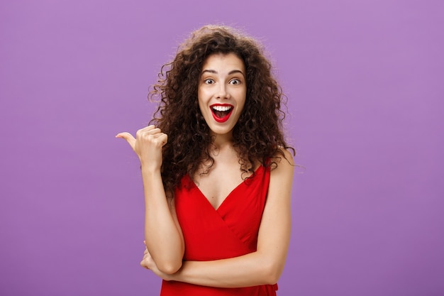 Energiegeladene attraktive und erstaunte gutaussehende Frau mit lockiger Frisur, die erstaunt lächelt, überrascht nach links zeigt mit dem Daumen, der Frage nach interessanter Anzeige in rotem Kleid über lila Wand stellt.