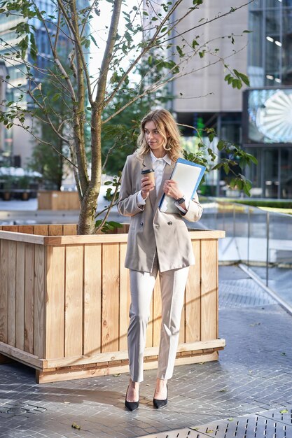 Energiegeladene Geschäftsfrau trinkt Kaffee zum Mitnehmen zum Mittagessen in der Nähe des Bürogebäudes, das auf der Straße steht