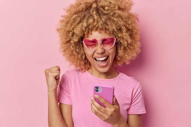 Energiegeladene, lockige Frau ballt die Faust, feiert den Erfolg, feiert den Sieg, hält das Smartphone, hat großartige Neuigkeiten, trägt eine herzförmige Sonnenbrille, ein lässiges T-Shirt, das über einem rosa Hintergrund isoliert ist. Ja, ich habe es geschafft