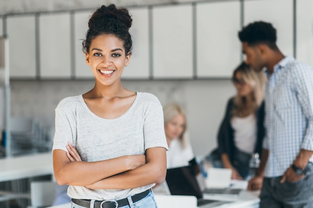 Enthusiastische afrikanische Studentin, die Gesellschaft mit Freunden im Hörsaal genießt. Innenporträt des lächelnden schwarzen Büroangestellten, der mit verschränkten Armen vor ausländischen Kollegen aufwirft.