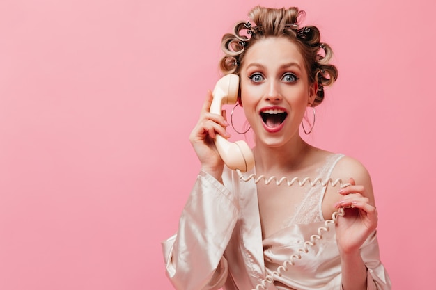 Enthusiastische Frau im rosa Gewand, die glücklich am Telefon spricht und auf isolierter Wand aufwirft