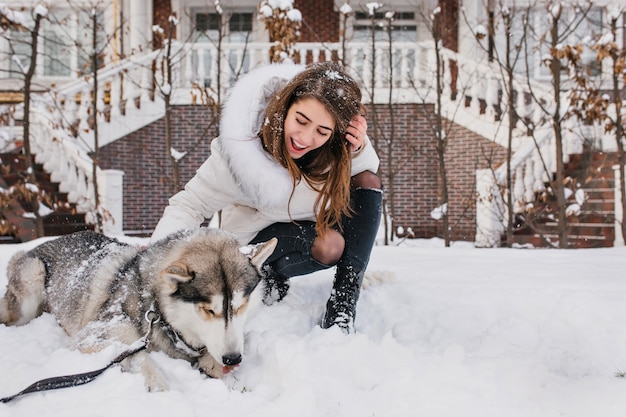 Enthusiastische Frau mit hellbraunem Haar, das ihren heiseren Welpen betrachtet und lächelt. Außenporträt der glückseligen jungen Frau, die mit Hund auf Schnee aufwirft.