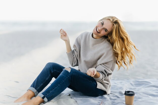 Enthusiastische kaukasische Frau, die Glück am Herbsttag am Strand ausdrückt. Inspirierte junge Frau in Jeans, die in der Natur lächeln