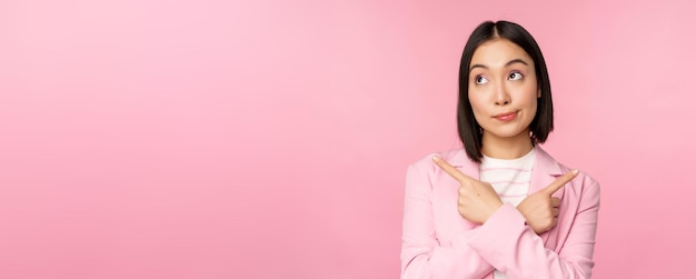 Entscheidungskonzept Junge asiatische Geschäftsfrau Corporate Worker zeigt mit dem Finger seitlich in zwei Richtungen und zeigt Varianten, die bei der Auswahl des rosafarbenen Hintergrunds unentschlossen aussehen