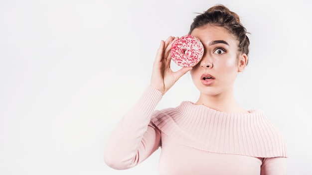 Entsetzte junge Frau schmollt ihre Lippen ab, welche die Augen mit Donut auf weißem Hintergrund bedecken