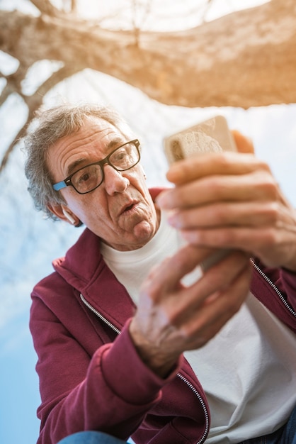 Entsetzter älterer Mann, der unter dem Baum betrachtet intelligentes Telefon sitzt