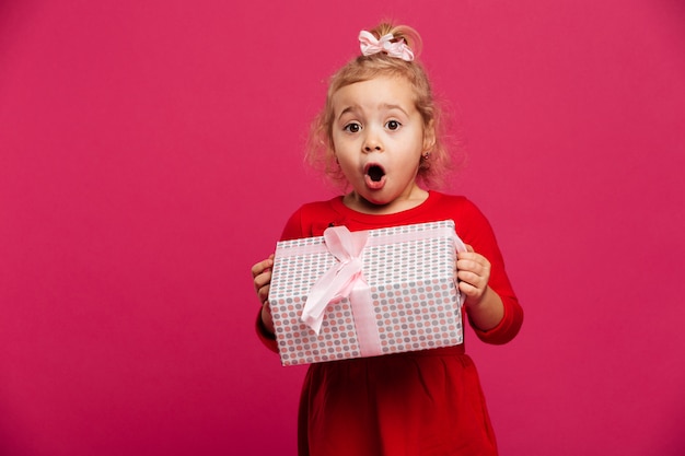 Entsetztes junges blondes Mädchen im roten Kleid, das Geschenkbox hält