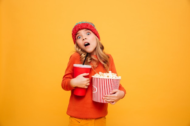 Entsetztes junges Mädchen in der Strickjacke und in Hut, die Popcorn- und Plastikschale beim Betrachten der Kamera über Orange halten