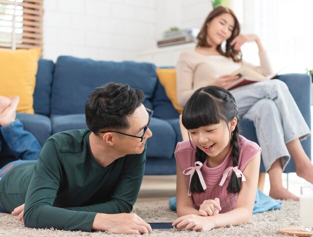 Entspannen Sie die asiatische Mutter, die ein Buch auf dem Sofa liest, und der Vater mit der Tochter, die Kunst im Wohnzimmer malt