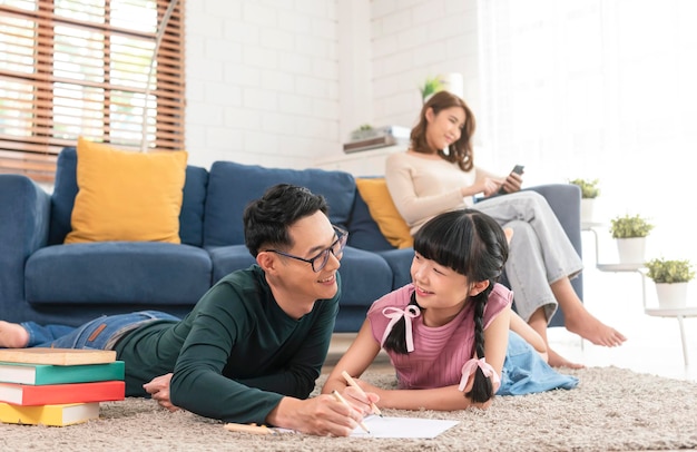 Entspannen Sie die asiatische Mutter, die ein Buch auf dem Sofa liest, und der Vater mit der Tochter, die Kunst im Wohnzimmer malt