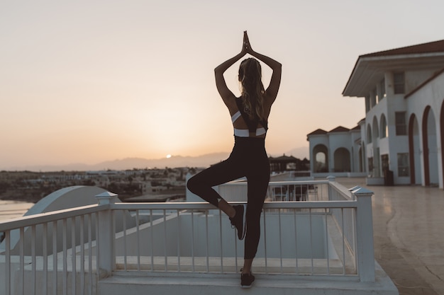 Entspannendes Yoga-Training der sportlichen attraktiven jungen Frau am Sonnenaufgang am Meer. Rückblick, Outwork, sportlicher Lebensstil, fröhliche Stimmung, Entspannung am Meer, Training im Freien, echte positive Emotionen.