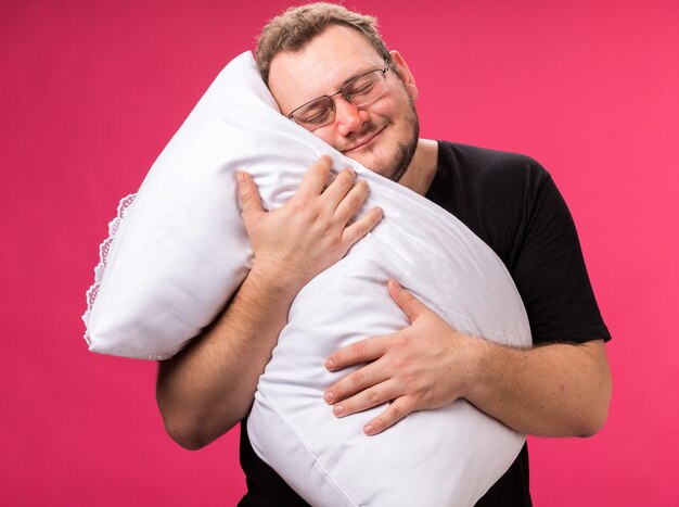 Entspannt mit geschlossenen Augen ein kranker Mann mittleren Alters umarmte Kissen isoliert auf rosa Wand