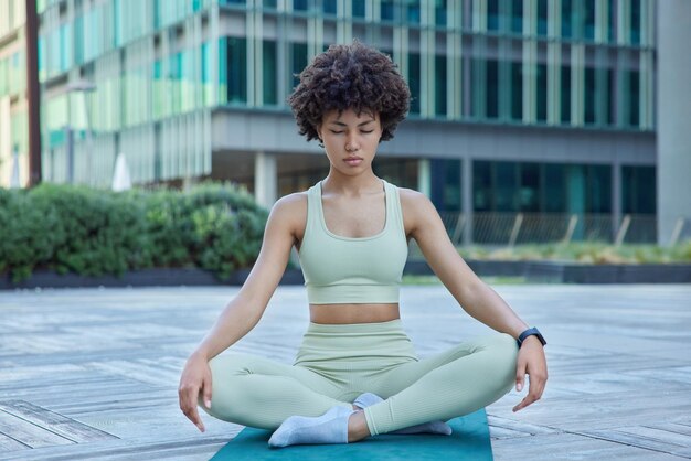 Entspannte, fitte Frau im Trainingsanzug sitzt in Lotus-Pose auf Karemat atmet tief ein, praktiziert Yoga, versucht, meditiert im Freien, posiert gegen modernes Stadtgebäude Gesundes Wohn- und Entspannungskonzept