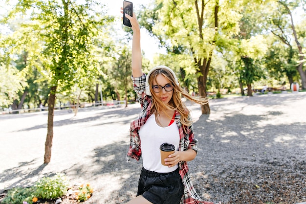 Entspannte Frau mit Tasse Kaffee lustiges Tanzen im Park. Debonair blonde Dame in Gläsern, die Musik auf Natur am Morgen genießen.