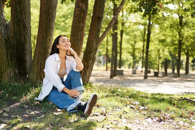 Entspannte junge Frau, die sich in der Nähe eines Baumes ausruht, der im Park auf dem Rasen unter Schatten sitzt, lächelt und glücklich aussieht