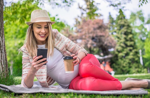 Entspannte junge Frau liegt auf einem Teppich auf dem Gras in einem Park oder Garten lächelnd und liest ihre SMS-Nachrichten auf ihrem Handy. Ganzkörperportrait der glücklichen Frau, die im Gras liegt