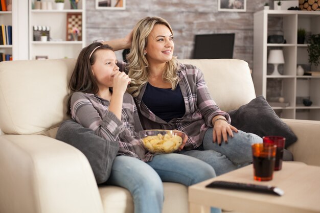 Entspannte junge Mutter und fröhliche Tochter vor dem Fernseher sitzen auf dem Sofa und essen Chips.