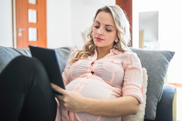 Entspannte junge schwangere Frau, die digitale Tablette verwendet