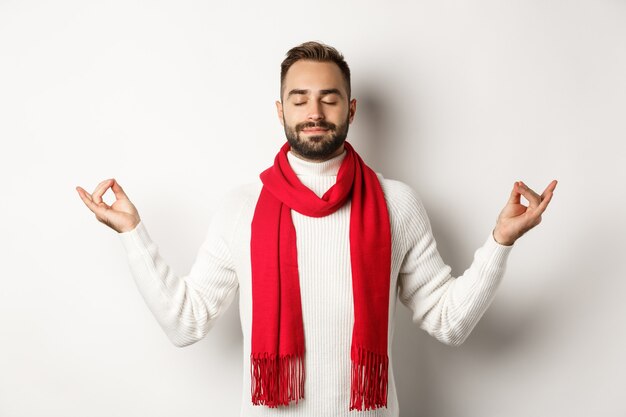 Entspannter bärtiger Kerl, der in Frieden steht, mit geschlossenen Augen meditiert, auf weißem Hintergrund in rotem Schal und Pullover steht