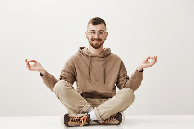 Entspannter fröhlicher Hipster sitzt auf dem Boden und meditiert, bleibt ruhig