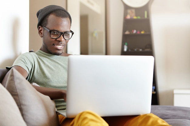 Entspannter fröhlicher junger schwarzer europäischer Mann in stilvoller Brille und Hut, die auf bequemem Sofa zu Hause mit Laptop-PC auf seinem Schoß sitzen, Videoanruf haben oder Videospiele online am Wochenende spielen