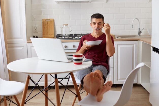 Entspannter, gutaussehender Mann mit kastanienbraunem Casual-T-Shirt, der am Tisch sitzt, die Füße auf den Stuhl setzt, Suppe isst und Film sieht, am Wochenende frühstückt oder eine Pause von der Online-Arbeit macht.
