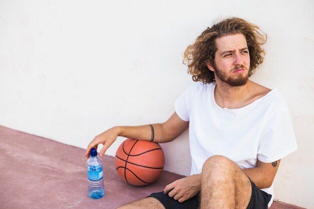 Entspannter junger Basketball-Spieler, der mit Ball- und Wasserflasche sitzt