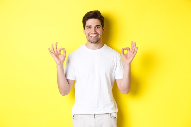 Entspannter Kerl lächelt, zeigt gute Zeichen, billigt oder stimmt zu, steht vor gelbem Hintergrund.